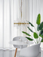Slimmer Clérachic chandelier on a chain with a few tall glass rods, hanging above a white round table beside a leafy plant and sheer curtains.