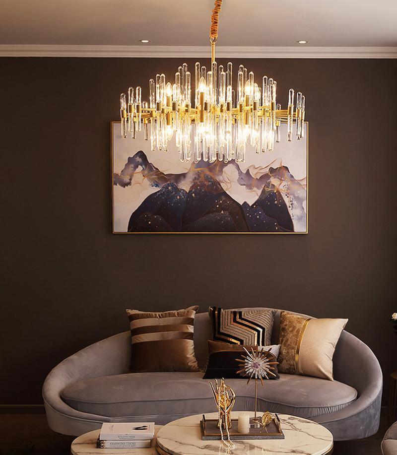 A Clérachic brass and glass rod chandelier lit above a grey curved sofa, with a mountain-print painting on a brown wall.