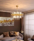 A Clérachic brass chandelier with clear glass rods glowing under a cream ceiling, above a sofa with patterned cushions and a golden painting.