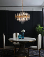 This Clérachic chandelier hangs above a round marble-top dining table with cream chairs, against a dark panelled wall and a tall plant.
