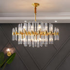 A Clérachic chandelier lit on a dark grey wall with diamond panelling, with a brass wall light and a leaf in the lower corner.