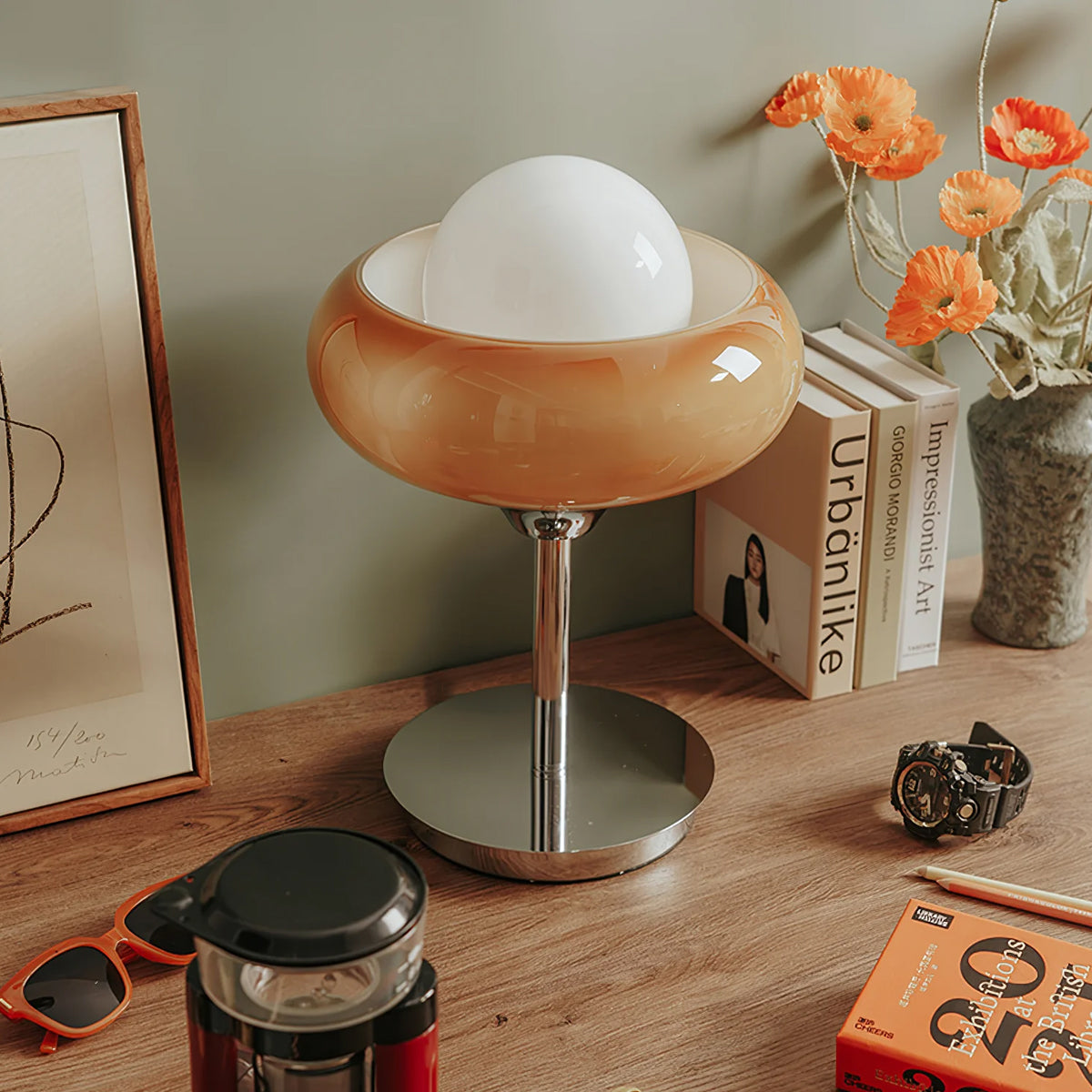 On a wooden desk with books, sunglasses and orange flowers, this Clérachic table lamp shows its amber shade over a chrome stem.