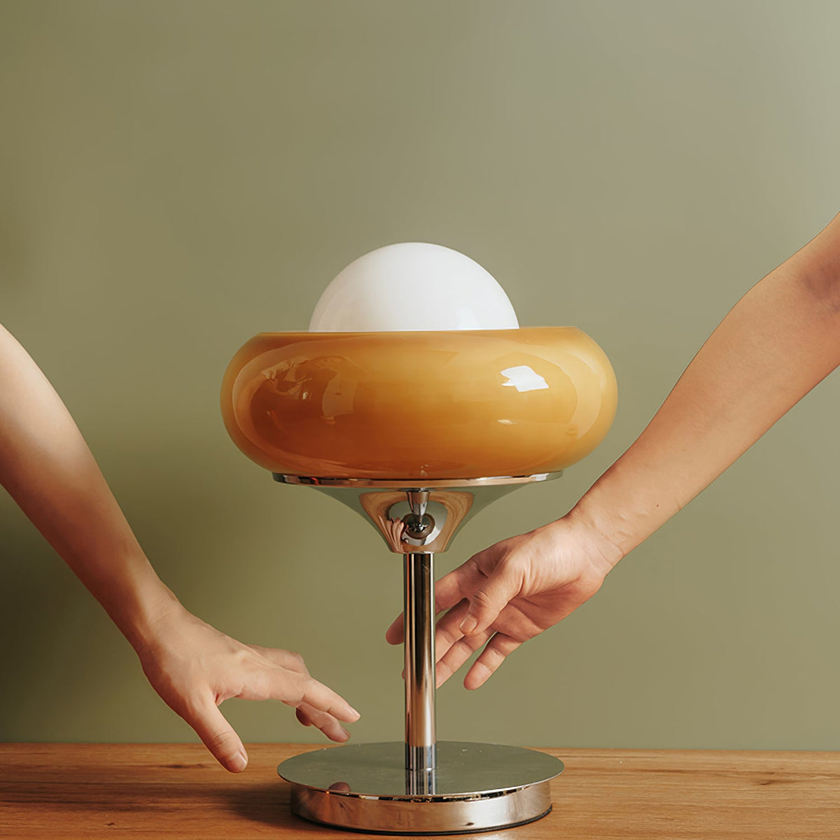 Two hands steady the chrome stem of this Clérachic table lamp on a wooden surface against a sage-green wall.