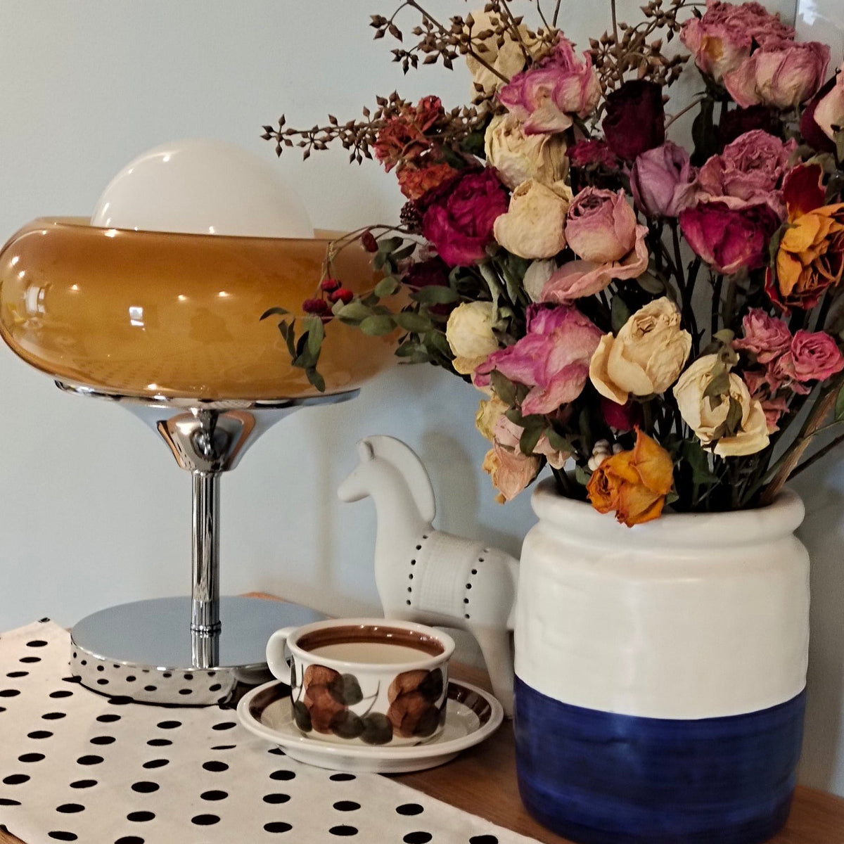 A Clérachic table lamp half hidden behind a bunch of dried roses in a blue and white jar, on a polka-dot cloth.