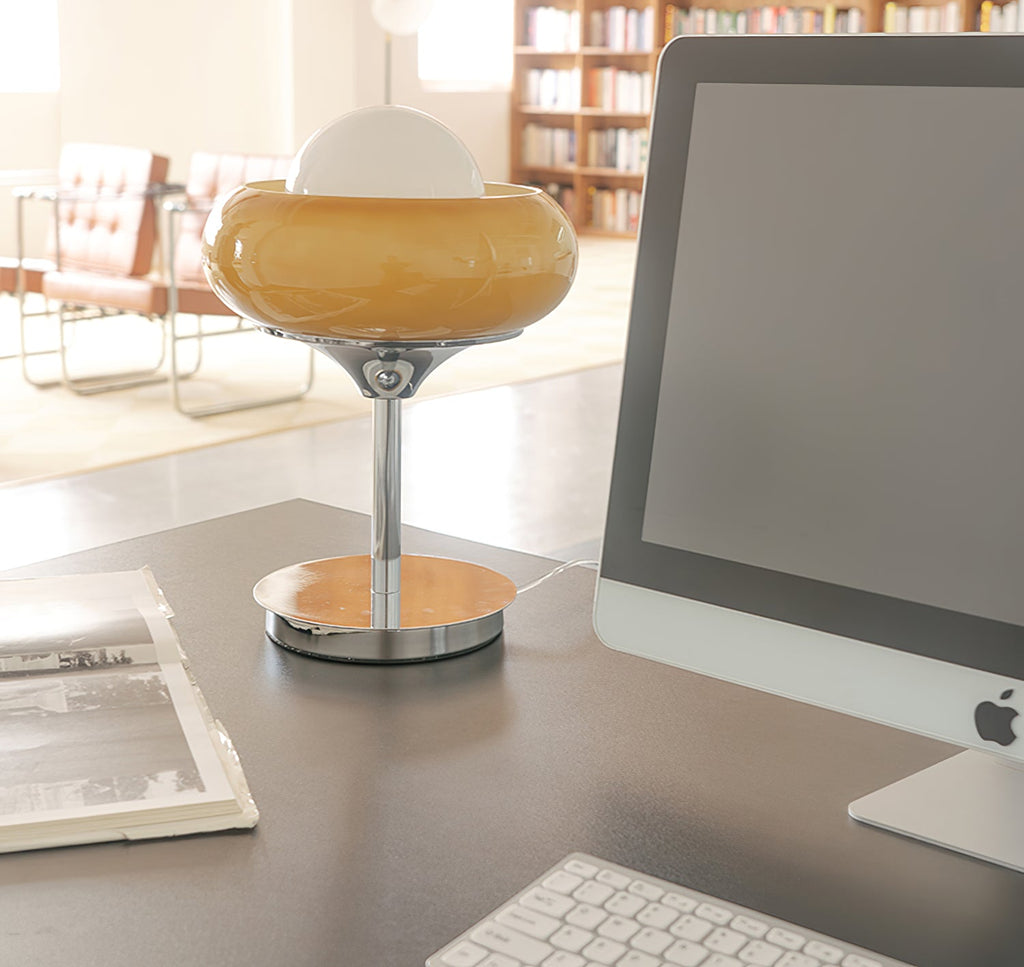 On a grey desk in front of a computer screen, this Clérachic table lamp stands with its amber shade and white globe unlit.