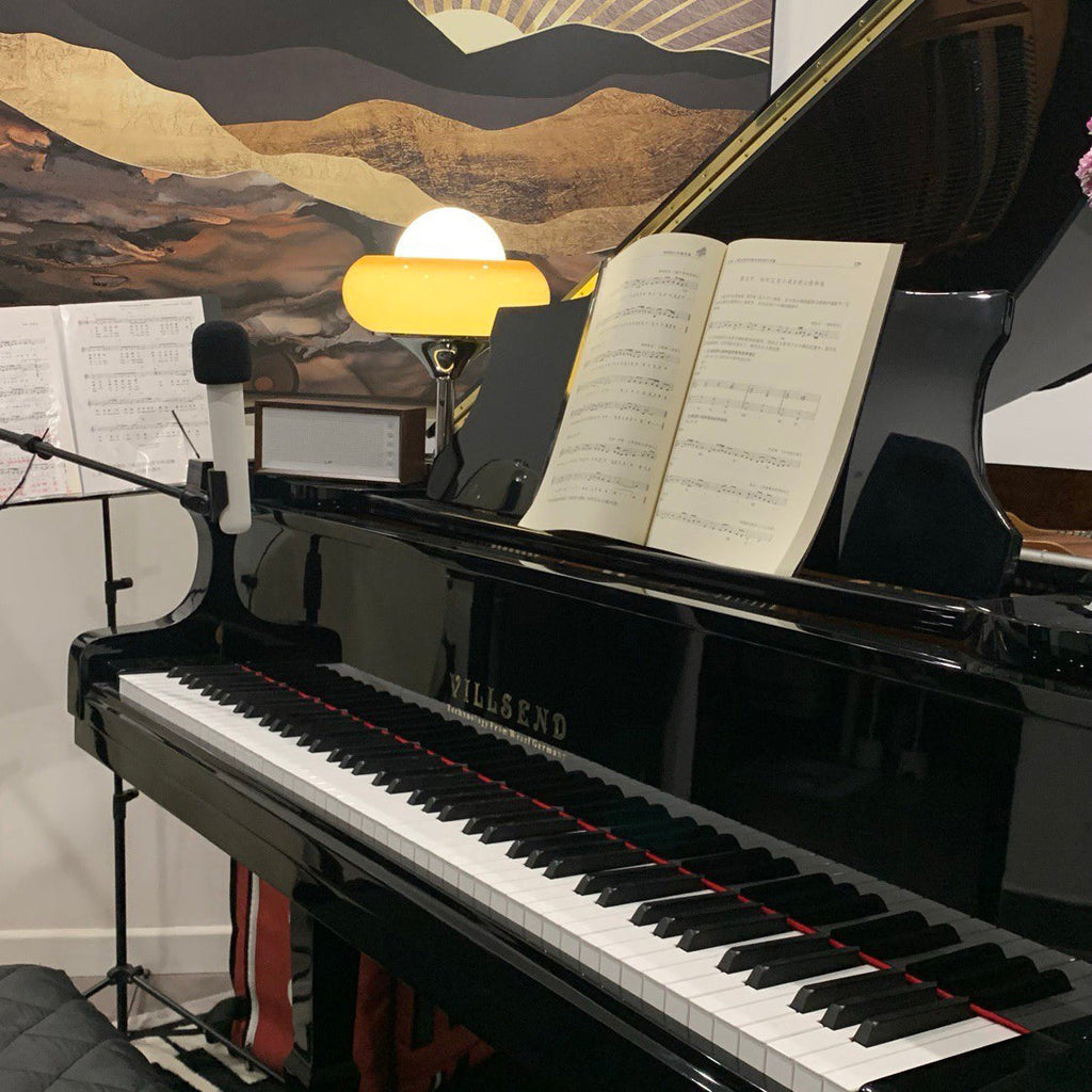 Small in the distance on top of a black piano, this Clérachic table lamp glows beside an open book of sheet music.