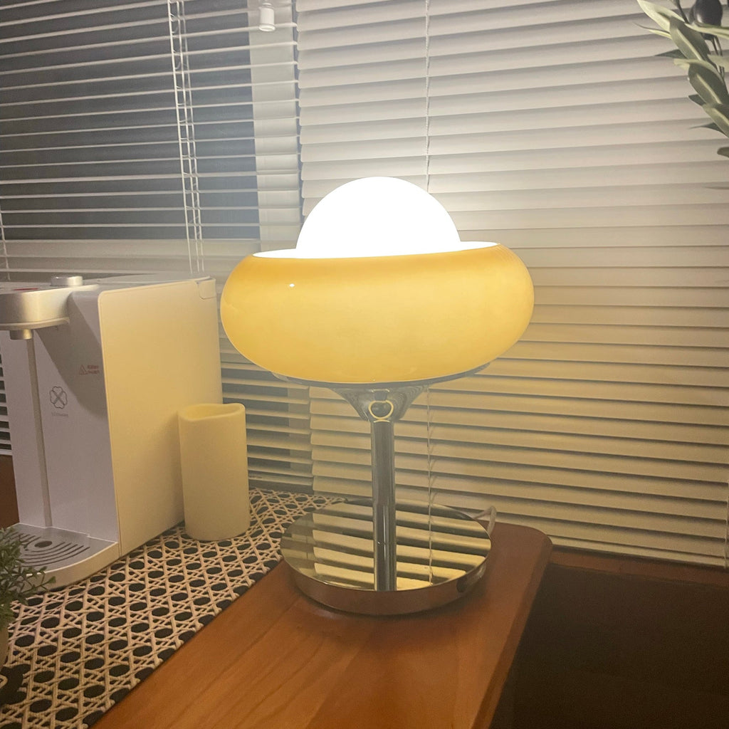 Lit warm in front of window blinds, this Clérachic table lamp stands on a wooden table next to a coffee machine.