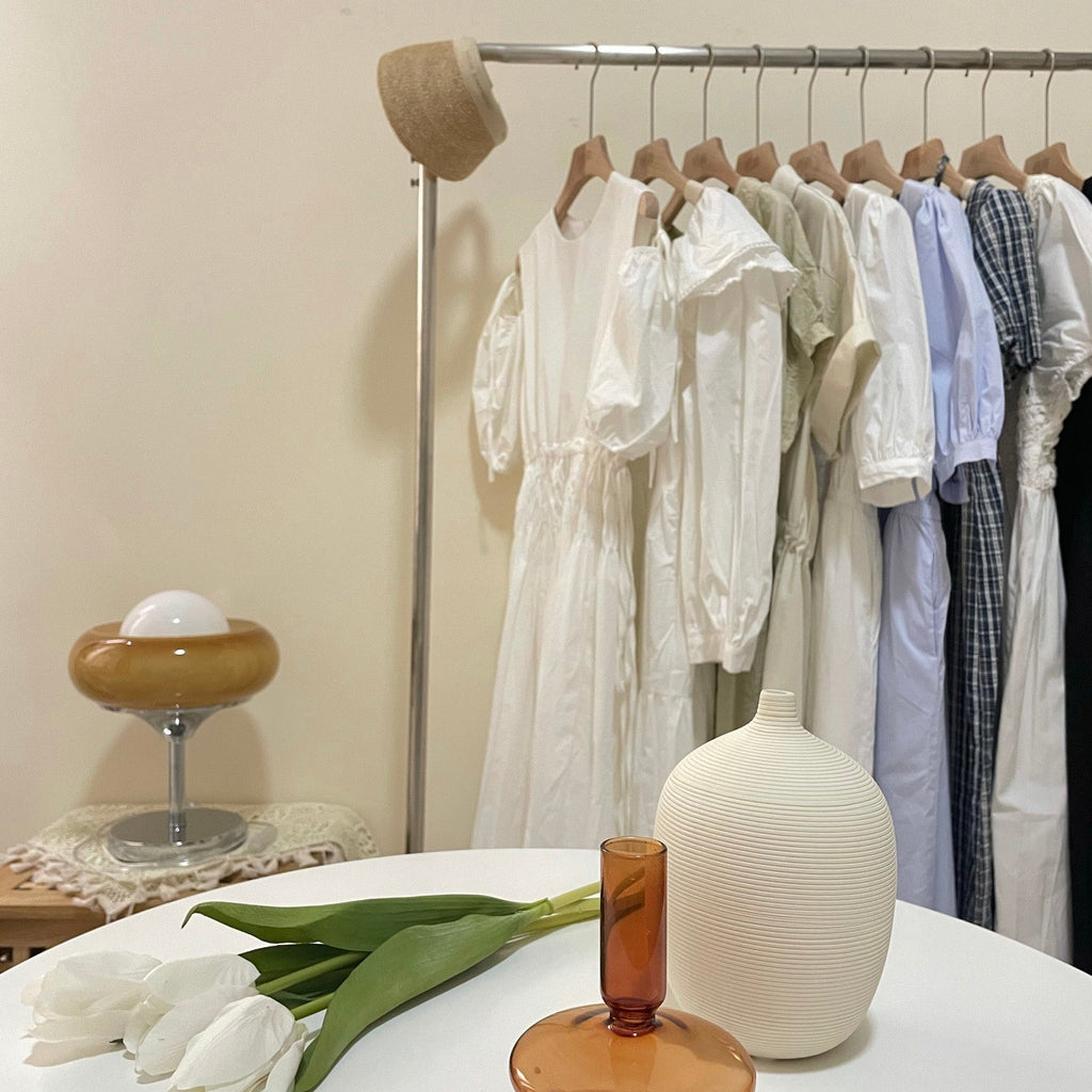 Small in a bedroom corner behind white tulips and a ribbed vase, this Clérachic table lamp sits near a rail of white dresses.