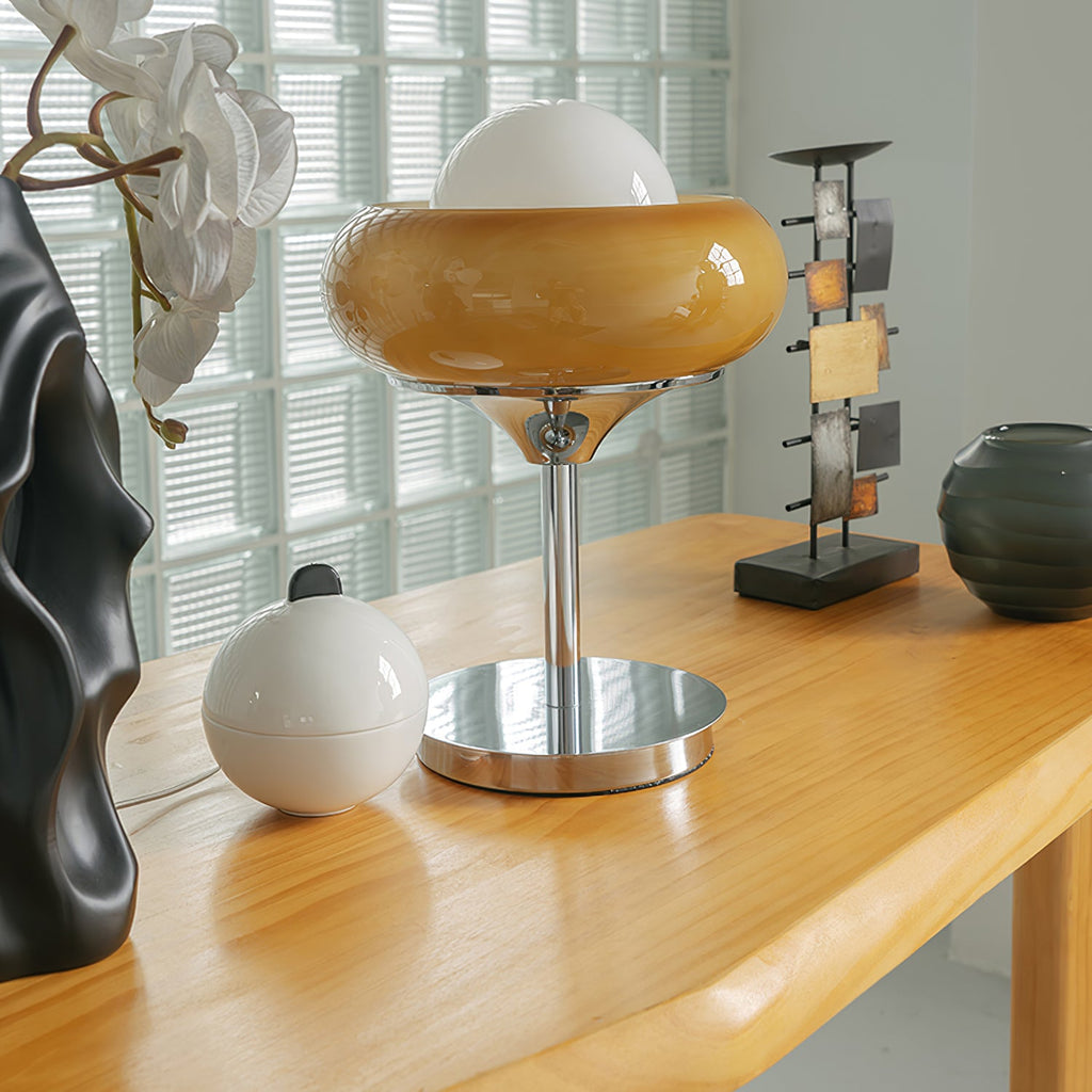On a light wood table beside a black sculpture and a metal shelf, this Clérachic table lamp shows its glossy amber shade.