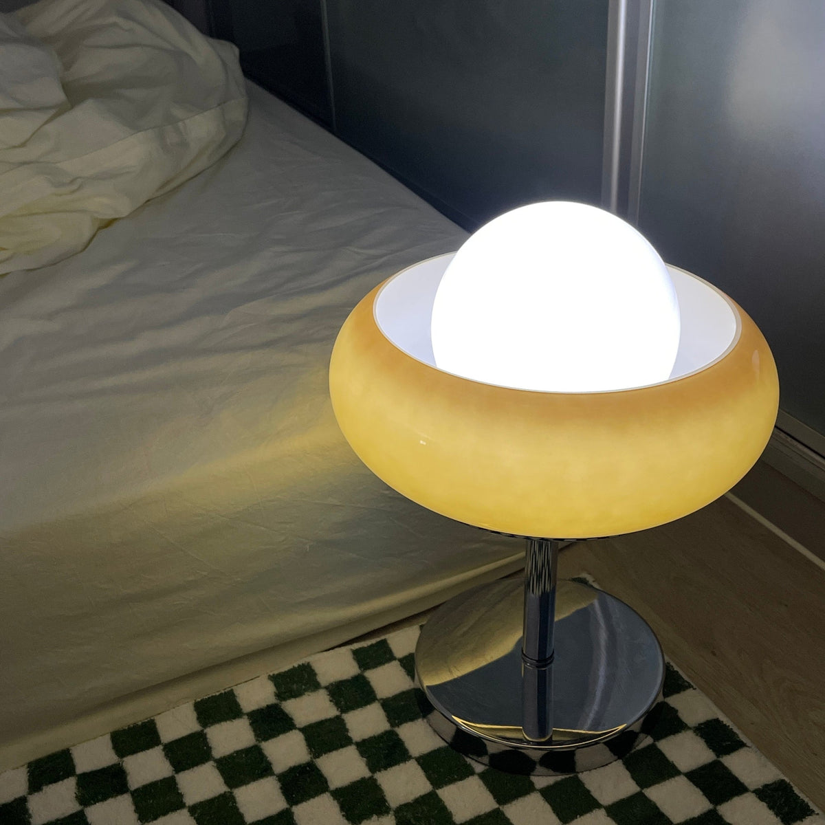 Close view of this Clérachic table lamp lit beside a bed, the white globe bright and the shade glowing soft yellow.