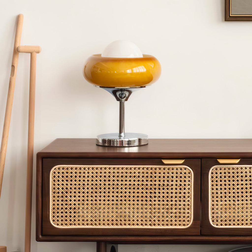 This Clérachic table lamp stands on a rattan-fronted wooden sideboard against a white wall, with a wooden stick leaning nearby.