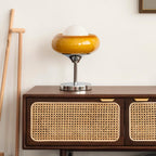 This Clérachic table lamp stands on a rattan-fronted wooden sideboard against a white wall, with a wooden stick leaning nearby.