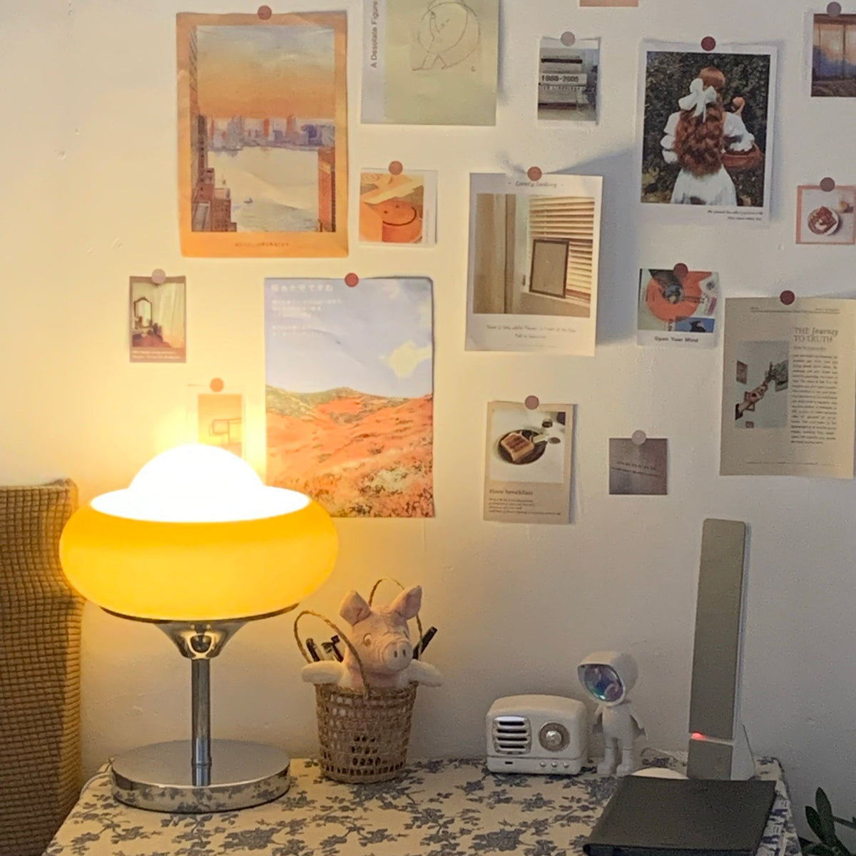 Lit above a desk of photo prints pinned to the wall, this Clérachic table lamp glows yellow beside a small radio and a plush toy.