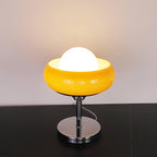 This Clérachic table lamp lit on a black surface, its white globe inside a bright yellow bowl shade, over a chrome base.