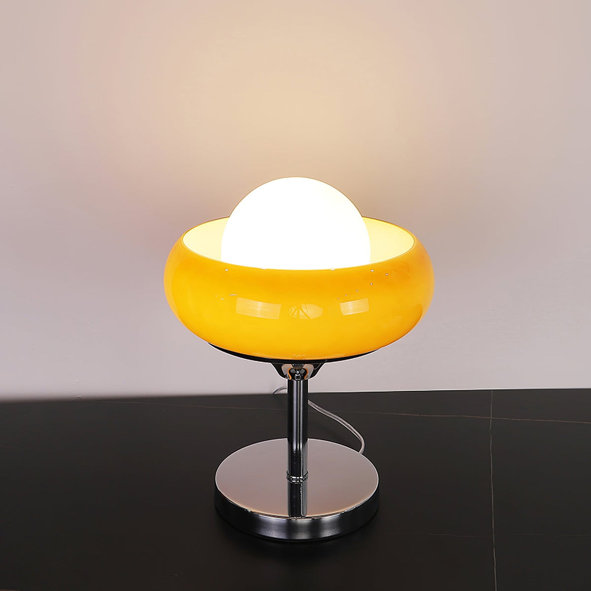 This Clérachic table lamp lit on a black surface, its white globe inside a bright yellow bowl shade, over a chrome base.