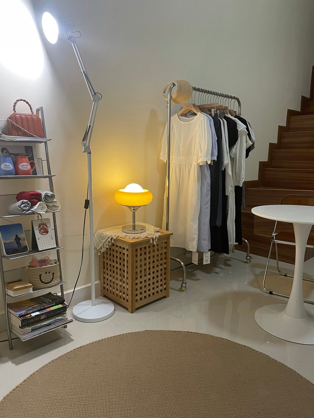 Small and lit in a room corner, this Clérachic table lamp sits on a woven box next to a clothes rail and a white round table.