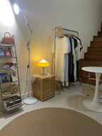 Small and lit in a room corner, this Clérachic table lamp sits on a woven box next to a clothes rail and a white round table.