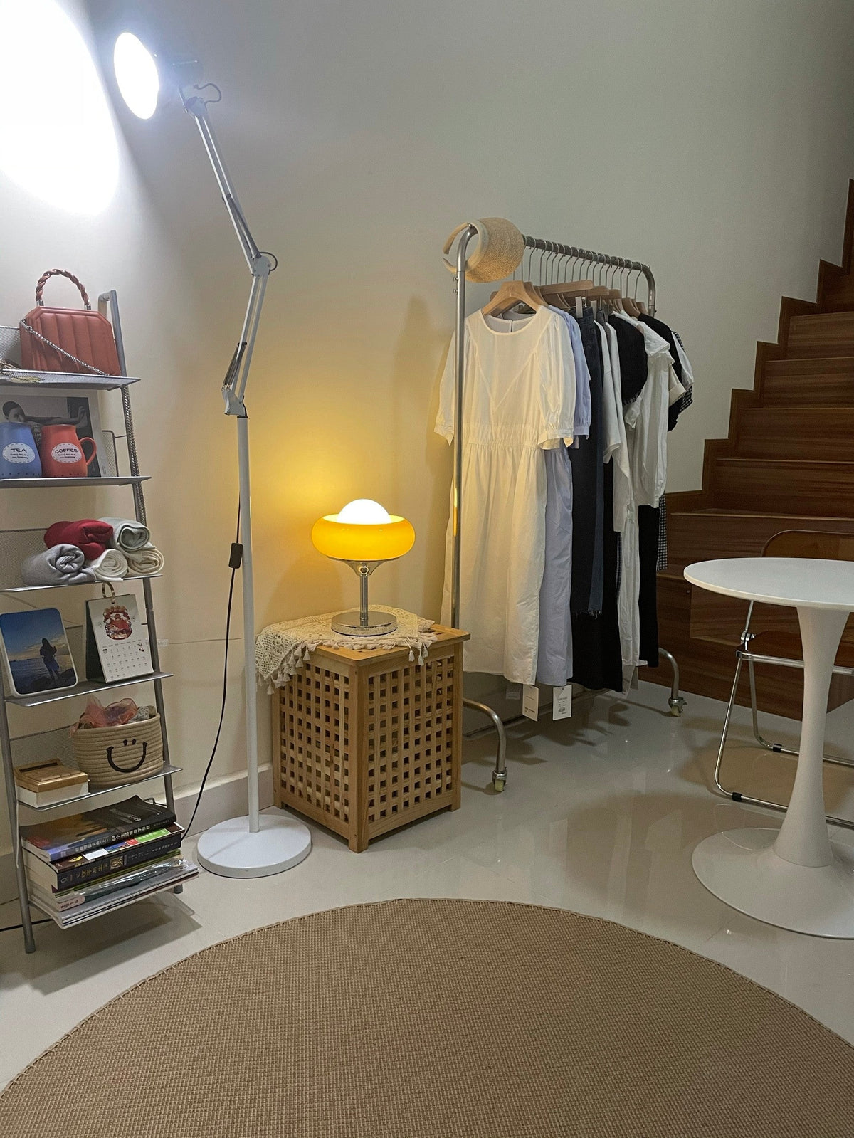 Small and lit in a room corner, this Clérachic table lamp sits on a woven box next to a clothes rail and a white round table.