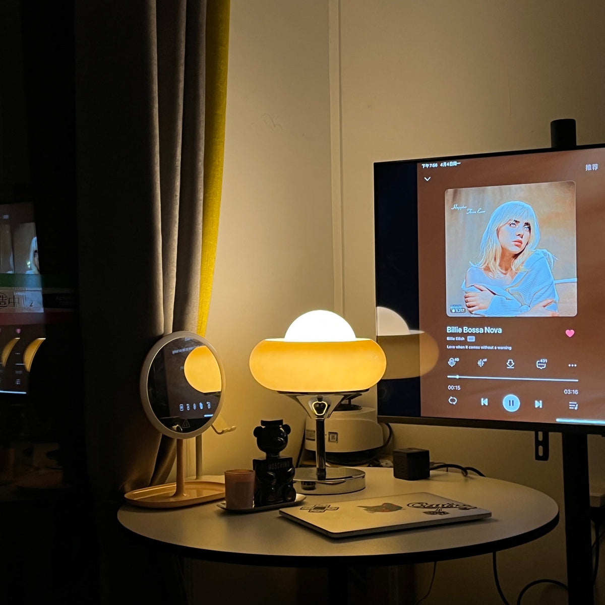 At night beside a monitor and a small round mirror, this Clérachic table lamp glows warm on a round desk.