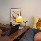 On a wooden desk with a laptop, small plants and a framed print, this Clérachic table lamp glows yellow.