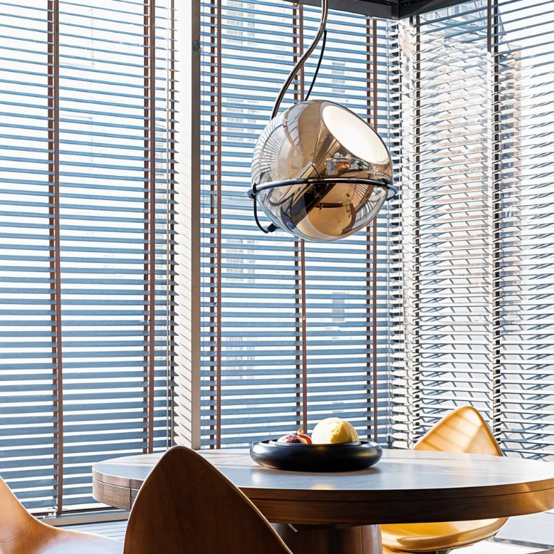 Clérachic amber glass pendant light hangs over a round dining table, its chrome arm catching light from slatted blinds.