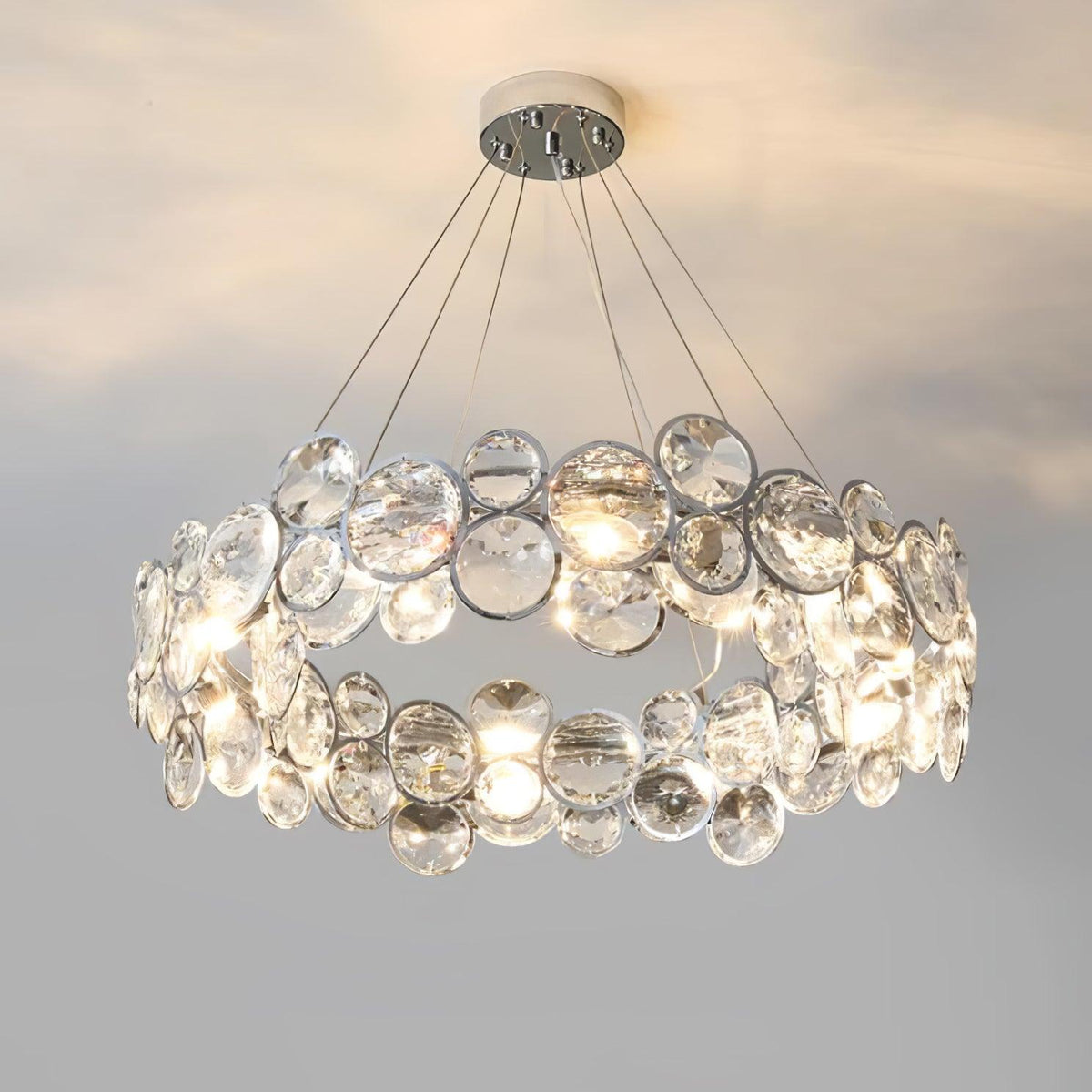 Round Clérachic crystal chandelier on a round chrome canopy, glowing warm against a pale grey wall.