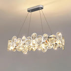 Oval Clérachic crystal chandelier lit and sparkling, hanging from a long chrome canopy against a grey wall.