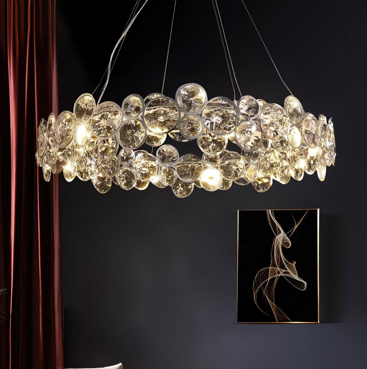 Wide view of a lit Clérachic crystal chandelier in a dark room, with a maroon curtain and a framed abstract print.