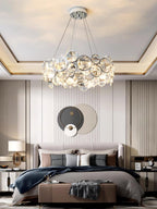 Round Clérachic crystal chandelier in a bedroom with a tray ceiling, a grey upholstered bed and a circular wall panel.