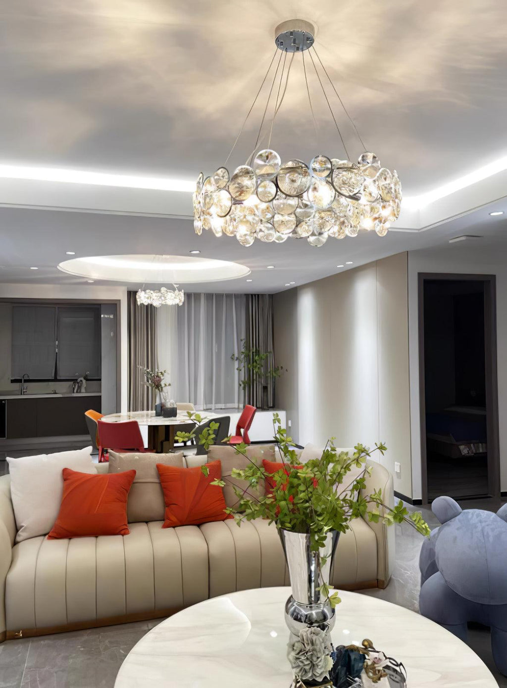 A Clérachic crystal chandelier lit in a living room, above a cream sofa with orange cushions and a white coffee table.