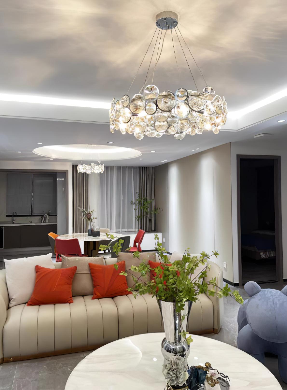 A Clérachic crystal chandelier lit in a living room, above a cream sofa with orange cushions and a white coffee table.