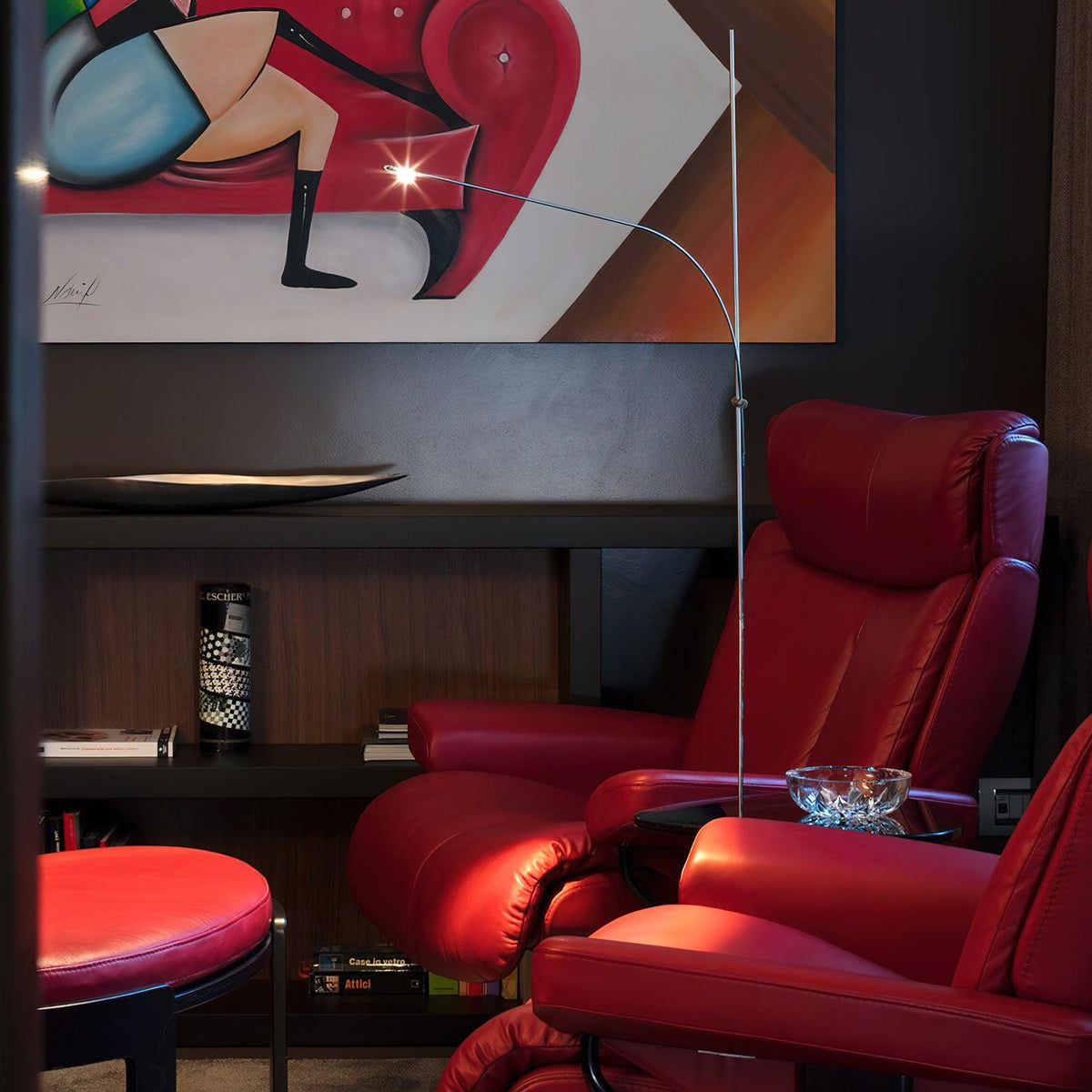 A Clérachic slender floor lamp arcs over a pair of red leather recliners, its small lamp head glowing in a dark-panelled room.