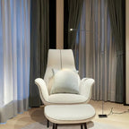 Beside a cream wingback armchair and footstool, this Clérachic chrome floor lamp curves out in front of grey-green curtains.