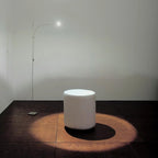 This Clérachic floor lamp throws a warm round pool of light across a dark wooden floor, lighting a white pouf in an empty room.