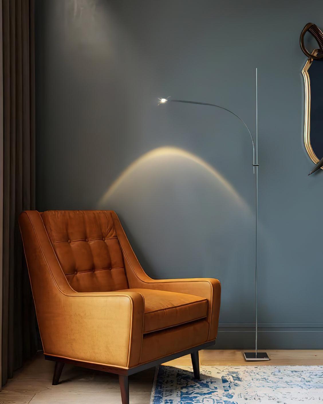 A Clérachic chrome floor lamp on a blue-grey wall, its beam arching above a tan leather armchair on a pale patterned rug.