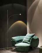 This Clérachic floor lamp stands behind a green velvet armchair, its warm beam rising in an arc up a charcoal wall.
