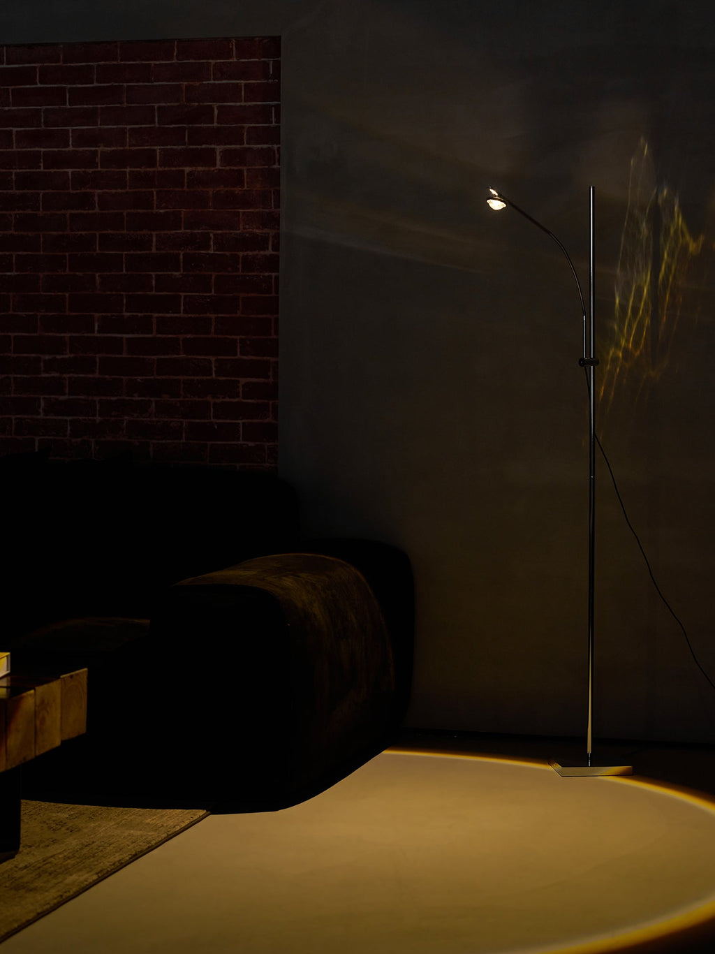 A Clérachic chrome floor lamp in a dim room with a brick wall, a faint glow on the floor beside a dark velvet sofa.