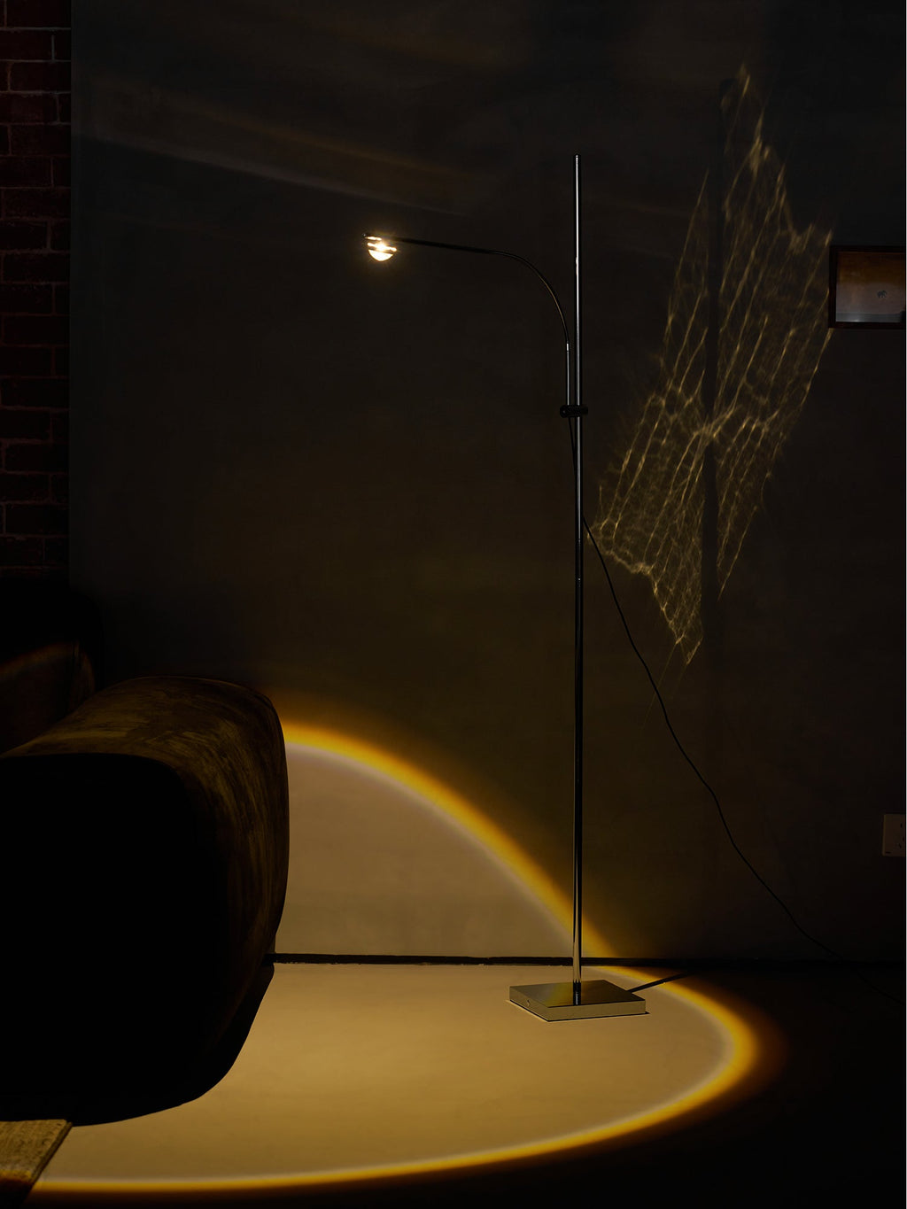 This Clérachic floor lamp lights a dim corner with a warm arc on the floor, beside a grey velvet sofa and a brick wall.