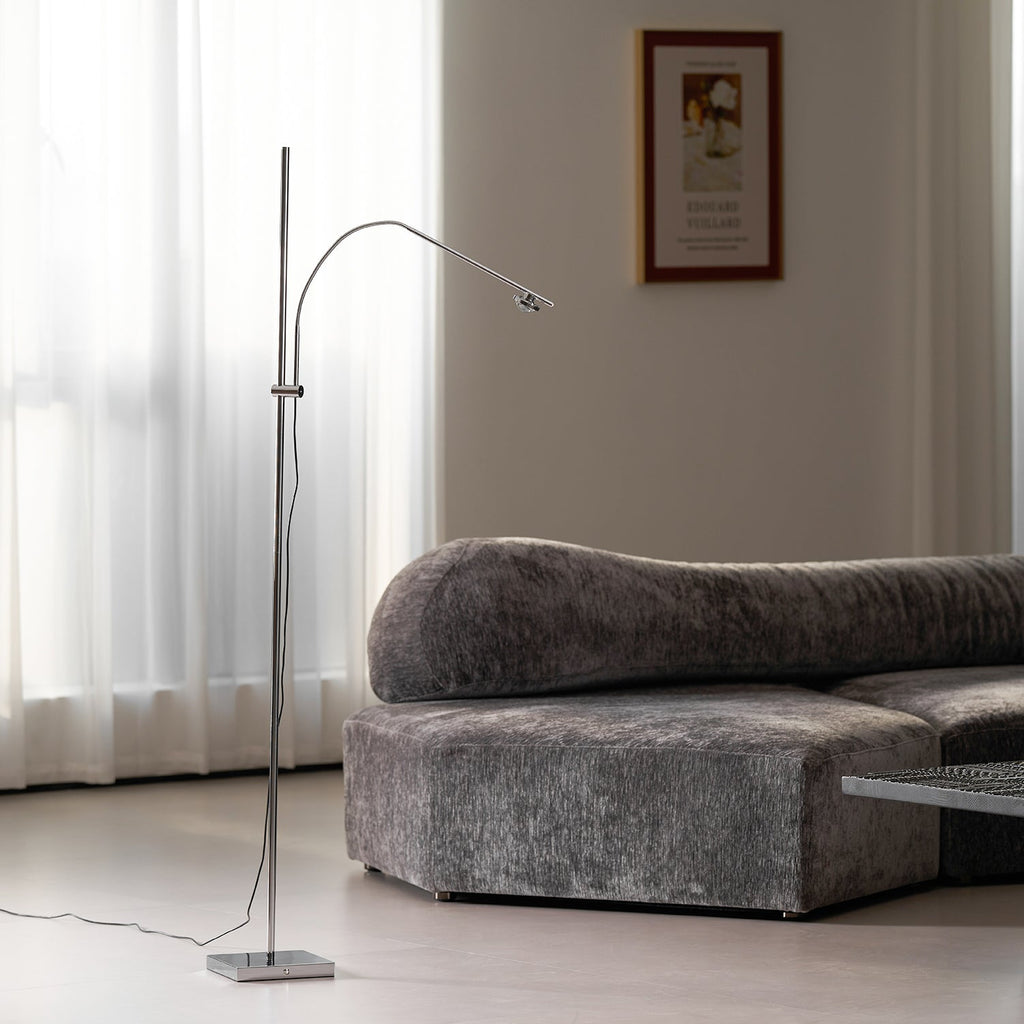 Beside a curvy grey velvet sofa and sheer curtains, this Clérachic chrome floor lamp reaches over with its lamp head angled down.
