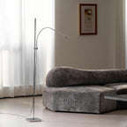 Beside a curvy grey velvet sofa and sheer curtains, this Clérachic chrome floor lamp reaches over with its lamp head angled down.