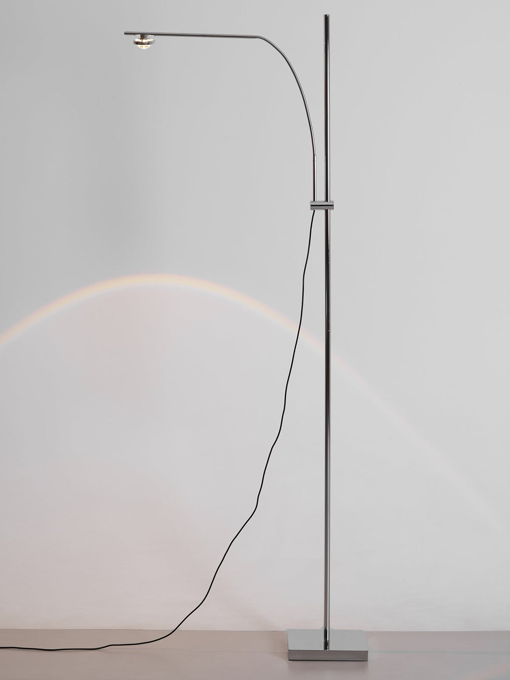 This Clérachic chrome floor lamp on a pale grey wall, with a faint arched glow of light and a cord looping to the base.