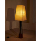 Lit Clérachic floor lamp glowing amber in a dim corner, its stitched shade casting a tall shadow on the wall beside it.