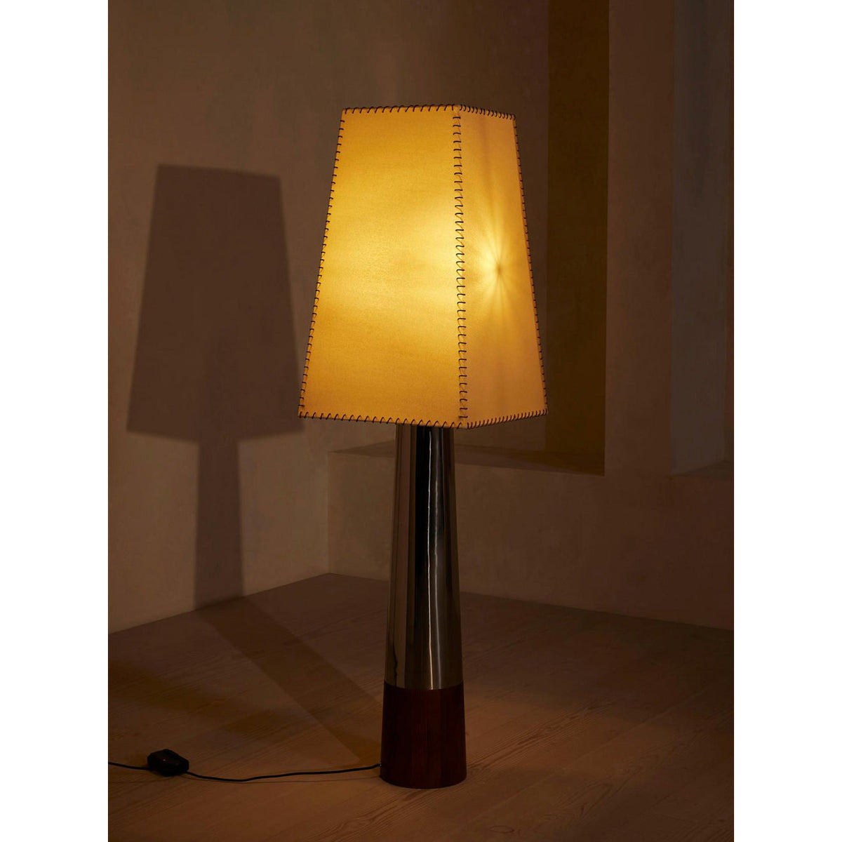 Lit Clérachic floor lamp glowing amber in a dim corner, its stitched shade casting a tall shadow on the wall beside it.