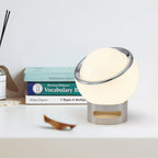 Clérachic globe table lamp glowing on a white shelf beside a stack of books and a fallen dry leaf, with a slotted chrome base.