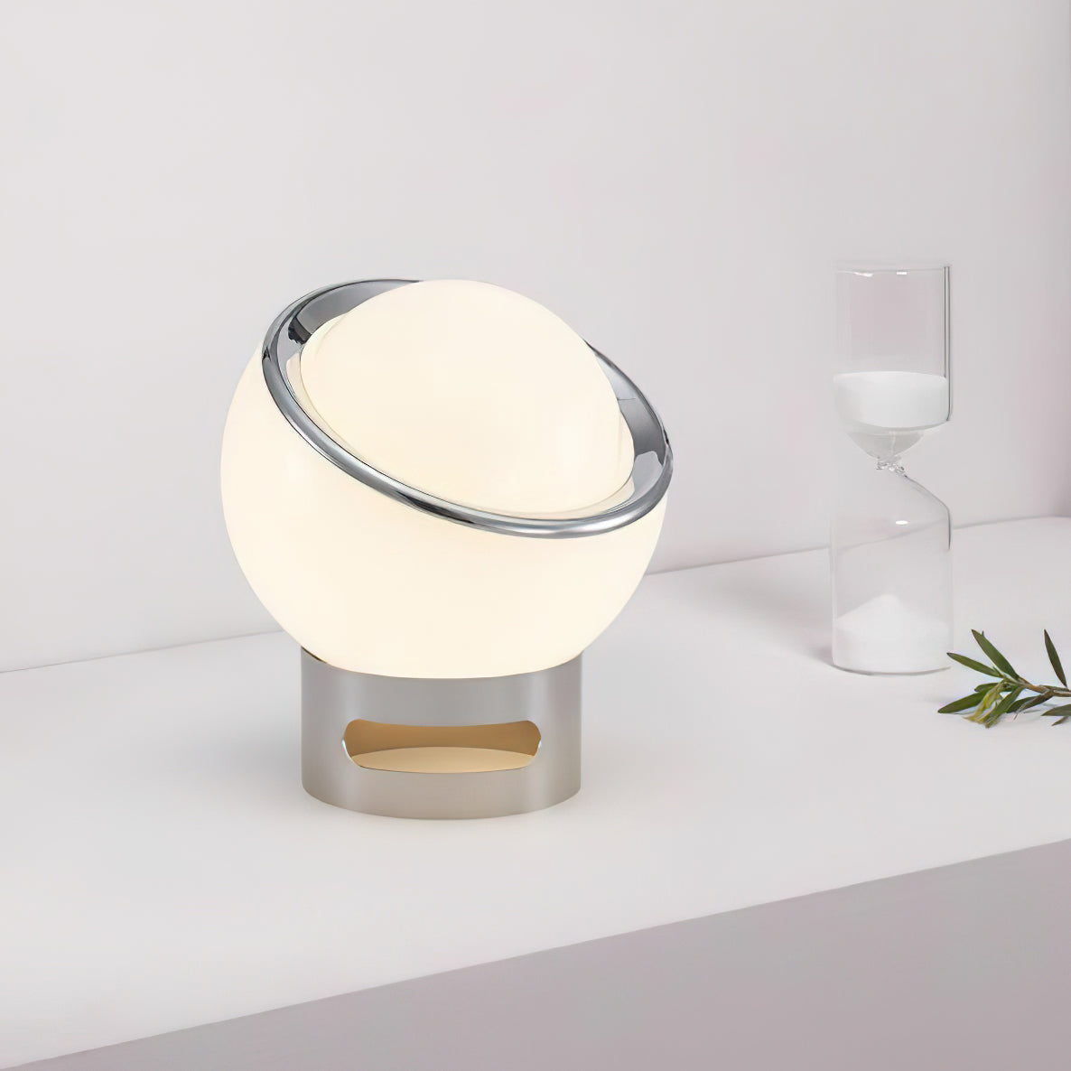 Soft light from the white glass globe of this Clérachic table lamp, on a pale desk next to an hourglass and a sprig of olive.