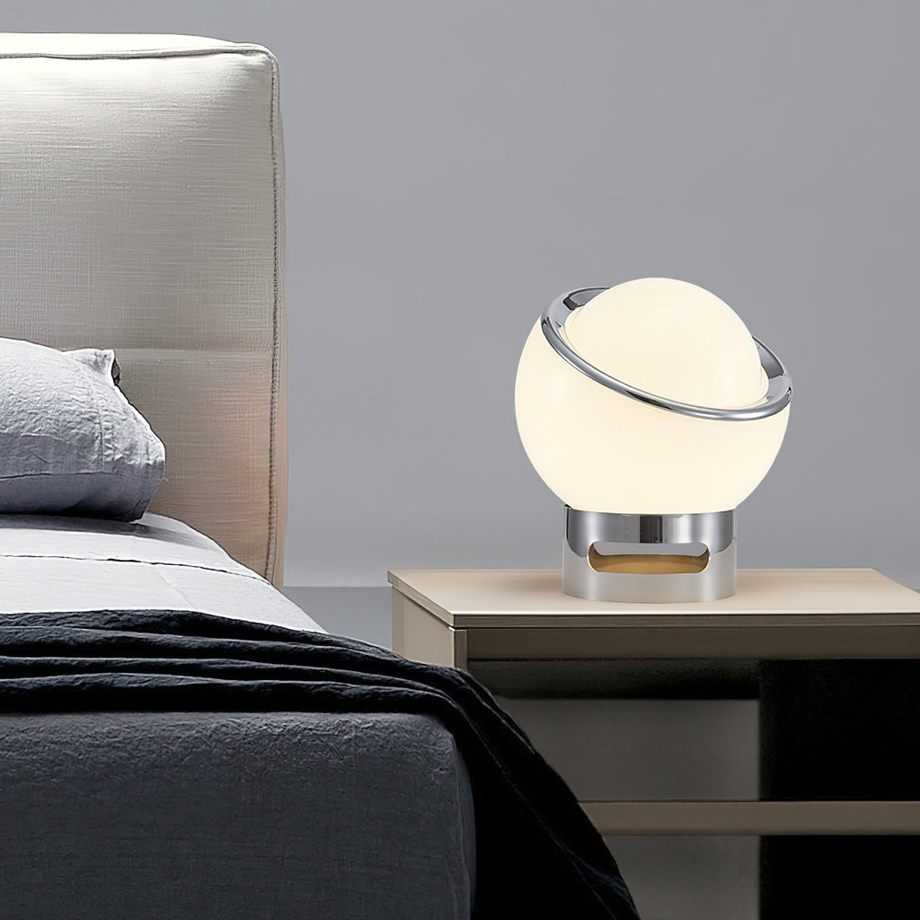 Small Clérachic globe table lamp lit on a pale wooden bedside table, with a grey headboard and dark bedding behind.