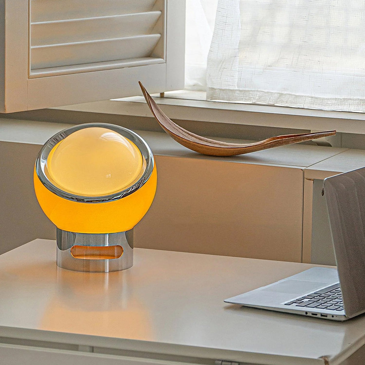 Orange-toned glass globe with a tilted chrome ring on this Clérachic table lamp, lit on a pale desk beside a laptop and window.
