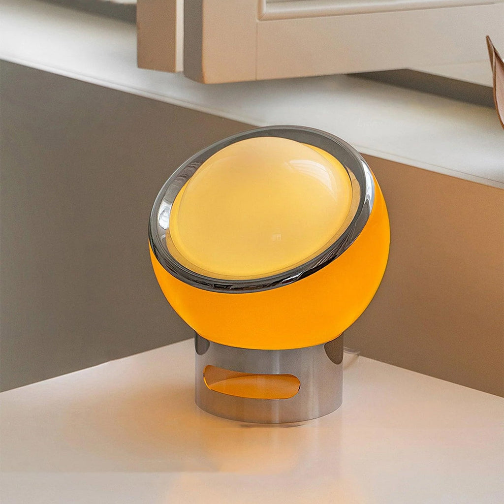 Angled view of an orange Clérachic table lamp lit on a white surface, its chrome ring framing the frosted glass dome.