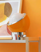 Orange version of this Clérachic table lamp against an orange wall and abstract art, with its chrome ring and base catching the light.