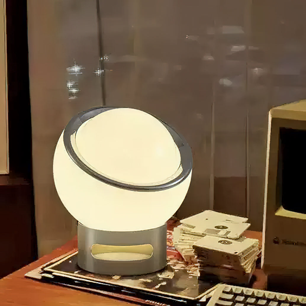 Large lit white globe on a Clérachic table lamp with a slotted chrome base, standing on a desk with old floppy disks and a computer.